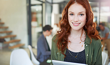 Eine junge Frau mit langen roten Haaren schaut lächelnd in die Kamera. In der Hand hält sie ein Tablet.