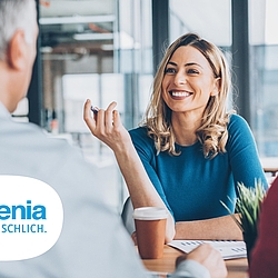 Eine blonde Frau sitzt an einem Tisch. Sie trägt einen blauen Pullover und lächelt. Ihr gegenüber sitzen Kunden. 