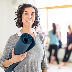 Eine Frau mit braunen Haaren steht in einem hellen Raum. Sie hält eine Yogamatte unter ihrem Arm und lächelt.