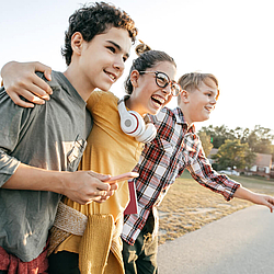 Zwei Jungen und ein Mädchen im Teenageralter umarmen sich und laufen eine Straße entlang. Der Junge im Vordergrund hält ein Smartphone. Das Mädchen in der Mitte lacht ausgelassen und trägt große Kopfhörer um den Hals.