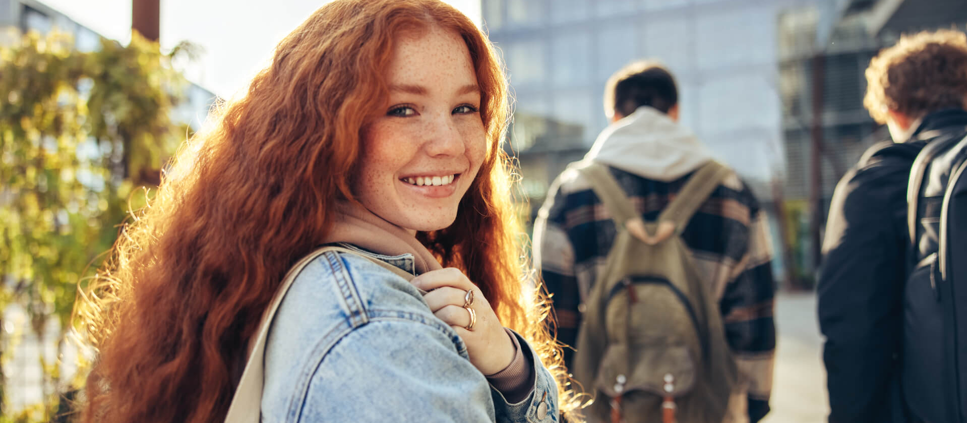 Eine junge Frau mit langen lockigen Haaren lacht in die Kamera. Sie traägt eine Jeansjacke und eine beige Tasche. Im Hintergrund gehen junge Männer an ihr vorbei.