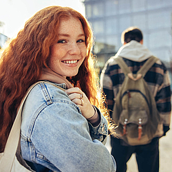 Eine junge Frau mit langen lockigen Haaren lacht in die Kamera. Sie trägt eine Jeansjacke und eine beige Tasche. 