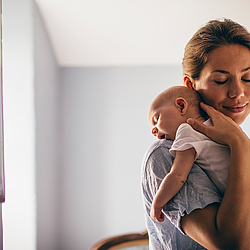 Ein junge Mutter hat ihr Neugeborenes auf ihre Schulter gelegt. Sie hat die Augen geschlossen und lächelt.