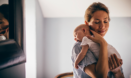 Ein junge Mutter hat ihr Neugeborenes auf ihre Schulter gelegt. Eine Hand hat sie am Rücken des Babys, die anderen unter seinem Po. Sie hat die Augen geschlossen und lächelt.