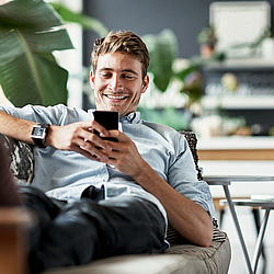 Ein junger Mann liegt auf dem Sofa und schaut auf sein Smartphone. Dabei lächelt er. 