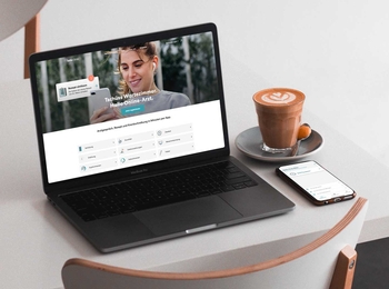 Auf einem Tisch stehen ein Laptopn, eine Tasse Kaffee und ein Smartphone. Auf dem Bildschirm des Laptops sieht man die Startseite der Teleclinic.
