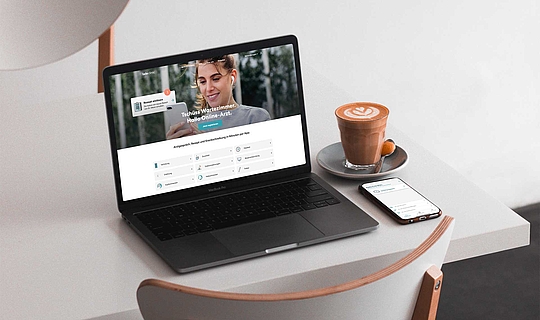 Auf einem Tisch stehen ein Laptopn, eine Tasse Kaffee und ein Smartphone. Auf dem Bildschirm des Laptops sieht man die Startseite der Teleclinic.