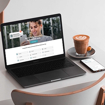 Auf einem Tisch stehen ein Laptopn, eine Tasse Kaffee und ein Smartphone. Auf dem Bildschirm des Laptops sieht man die Startseite der Teleclinic.