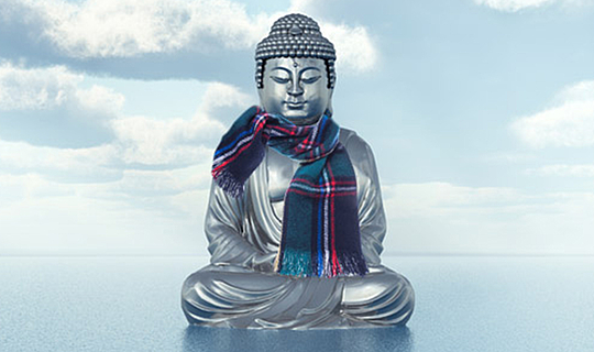 Eine Buddha-Statue. Sie steht auf dem Wasser, im Hintergrund sieht man einen blauen Himmel voller Wolken. Sie Statue trägt einen karierten Schal.