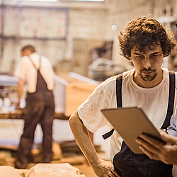 Ein Mann mit grauen Haaren schaut zusammen mit einem jungen Mitarbeiter auf ein Tablet. Im Hintergrund sieht man eine Werkstatt.