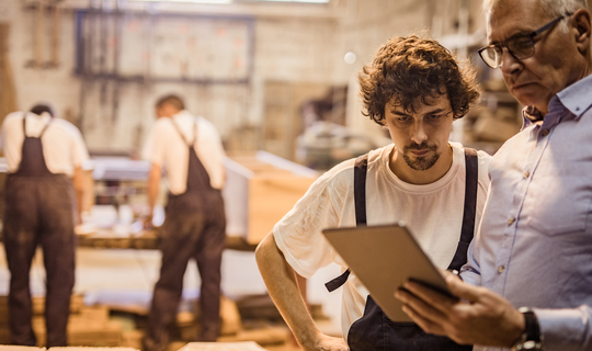 Ein ältere Mann mit grauen Haaren und Brille schaut zusammen mit einem jungen Mitarbeiter auf ein Tablet. Im Hintergrund sieht man eine Werkstatt.