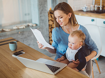 Eine Mutter sitzt mit ihrem Kind auf dem Schoß vor einem Laptop. Sie schaut sich einen Antrag an.