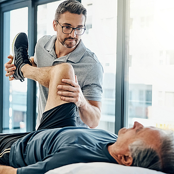 Ein älterer Mann liegt in Sportkleidung auf einer Behandlungsliege. Sein Therapeut hält während der Rehabilitationsübung sein Bein hoch. 