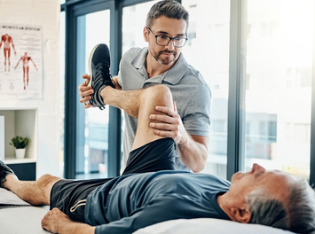 Ein älterer Mann liegt in Sportkleidung auf einer Behandlungsliege. Sein Therapeut hält während der Rehabilitationsübung sein Bein hoch. 