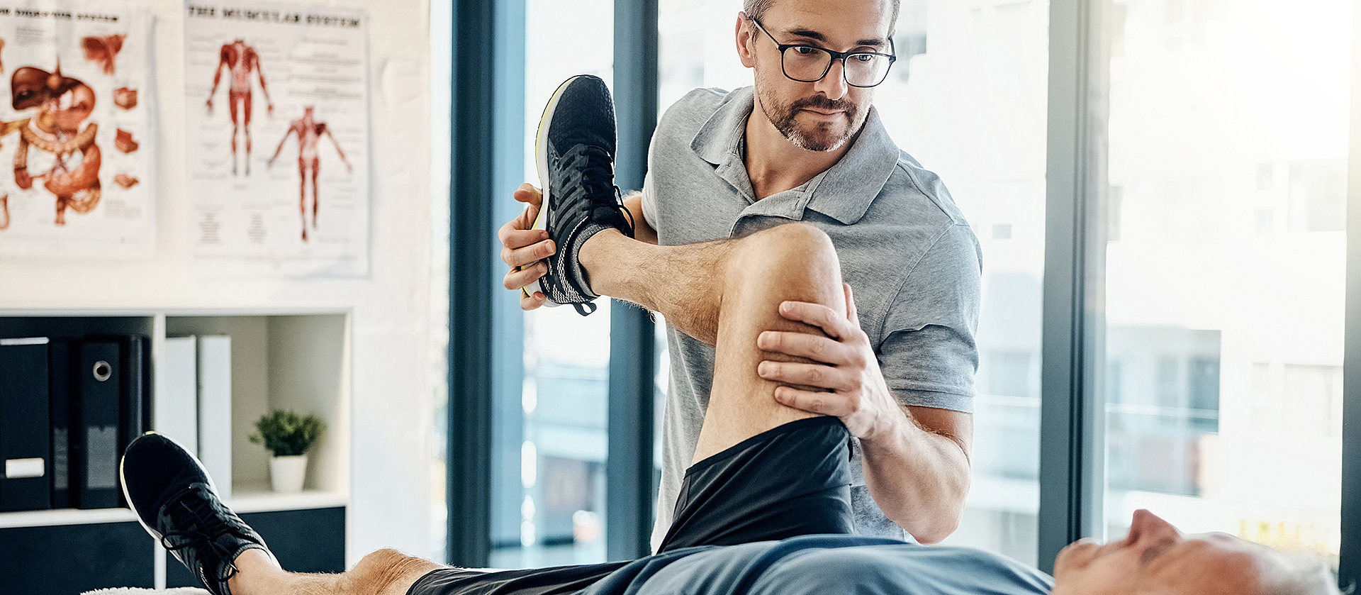 Ein älterer Mann liegt in Sportkleidung auf einer Behandlungsliege. Sein Therapeut hält während der Rehabilitationsübung sein Bein hoch. 