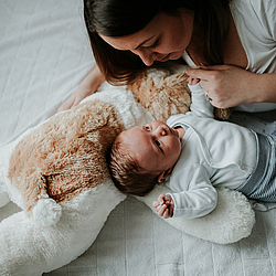 Eine junge Mutter liegt mit ihrem Säugling auf dem Bett. Die beiden schauen sich liebevoll an. Der Kopf des Babys liegt auf einem großen weichen Stofftier.