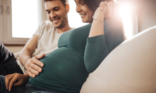 Eine junge schwangere Frau sitzt auf dem Sofa. Sie schaut lächelnd auf ihren Bauch. Ihr Partner hat den Arm um sie gelegt, seine andere Hand liegt auf ihrem Bauch.