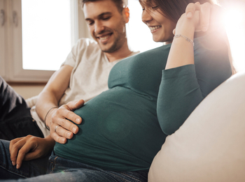 Eine junge schwangere Frau sitzt auf dem Sofa. Sie schaut lächelnd auf ihren Bauch. Ihr Partner hat den Arm um sie gelegt, seine andere Hand liegt auf ihrem Bauch.