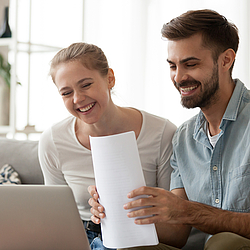 Ein junges Paar sitzt gemeinsam auf dem Sofa und schauen lachend auf einen weißen Laptop. Der Mann hält Unterlagen in der Hand.