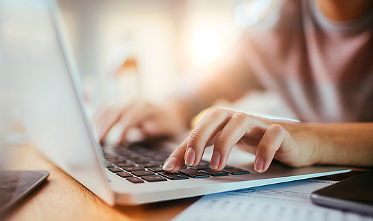 Eine Person tippt auf der Tastatur ihres grauen Laptops. Neben ihr liegen Zettel und ein Handy.