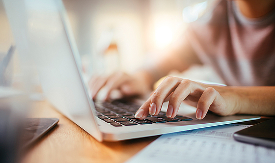 Eine junge Frau tippt auf der Tastatur ihres grauen Laptops. Neben ihr liegen Zettel und ein Handy.