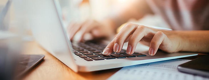 Eine junge Frau tippt auf der Tastatur ihres grauen Laptops. Neben ihr liegen Zettel und ein Handy.