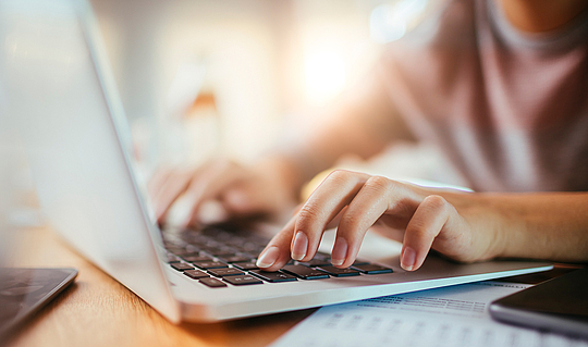 Eine junge Frau tippt auf der Tastatur ihres grauen Laptops. Neben ihr liegen Zettel und ein Handy.