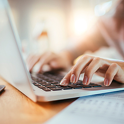 Eine Person tippt auf der Tastatur ihres grauen Laptops. Neben ihr liegen Zettel und ein Handy.