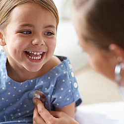Ein kleines Mädchen in einem blauen Sommerkleid lacht eine Ärztin an, die mit einem Stethoskop seine Brust abhört.