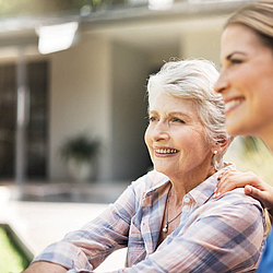 Eine ältere Frau sitzt zusammen mit einer Pflegerin im Garten. Sie Sonne scheint und sich lächeln.