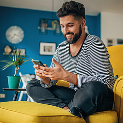 Ein Mann sitzt im Schneidersitz auf einem Sessel. Er schaut dabei auf sein Smartphone und lächelt.