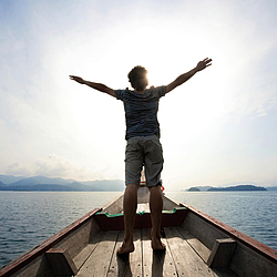 Ein Mann in sommerlicher Kleidung steht auf einem kleinen Boot auf dem Meer. Er hat die Arme ausgestreckt und genießt die Sonne.