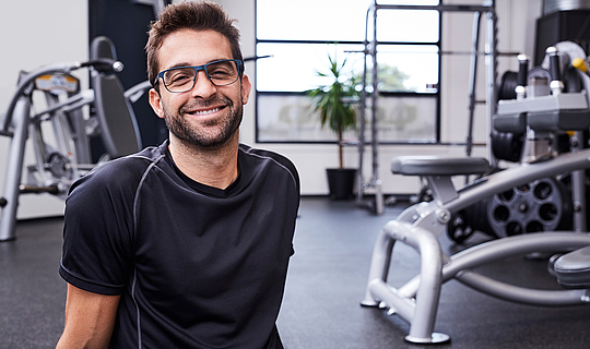 Ein dunkelhaariger Mann mit Brille und Sportkleidung und sitzt in einem Fitnessstudio. Er lächelt in die Kamera. 