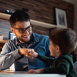Ein junger Vater mit dunklnen kurzen Haaren Bart und Brille, trägt ein blaues Jeanshemd. Er sitzt mit seinem Sohn an einem Tisch. Der kleine Junge hat kurze braune Haare und trägt einen grünen Pullover. Die beiden schauen sich lachend an und stoßen die Fäuste aneinander.