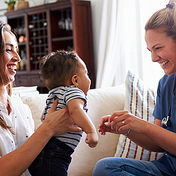 Eine junge Mutter mit langen Haaren hält ihr Baby einer lächelnden Hebamme entgegen. 