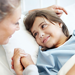 Ein junges Mädchen mit langen braunen Haaren trägt einen Patientenhemd und liegt in einem Krankenhausbett. Sie lächelt eine Frau an, die ihre Hand hält.