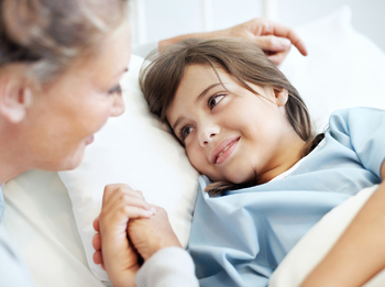 Ein junges Mädchen mit langen braunen Haaren trägt einen Patientenhemd und liegt in einem Krankenhausbett. Sie lächelt eine Frau an, die ihre Hand hält.
