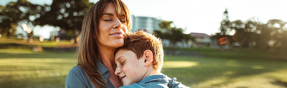 Eine Frau mit langen Haaren umarmt ihren Sohn. Sie stehen bei Sonnenschein auf einer Wiese.