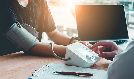 Ein Arzt sitzt mit einem Patienten an einem Tisch. Er misst den Blutdruck des Patienten. Neben ihm steht ein Laptop, auf dem Tisch liegt ein Klemmbrett mit einem Formular sowie ein Stift.
