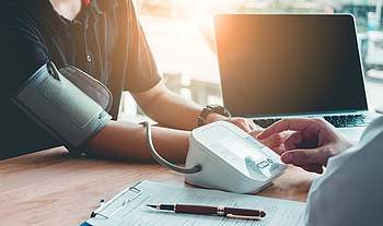 Ein Arzt sitzt mit einem Patienten an einem Tisch. Er misst den Blutdruck des Patienten. Neben ihm steht ein Laptop, auf dem Tisch liegt ein Klemmbrett mit einem Formular sowie ein Stift.