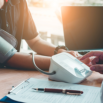 Ein Arzt sitzt mit einem Patienten an einem Tisch. Er misst den Blutdruck des Patienten. Neben ihm steht ein Laptop, auf dem Tisch liegt ein Klemmbrett mit einem Formular sowie ein Stift.