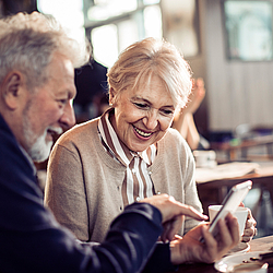 Ein älteres Paar mit sitzt in einem Café. Sie essen Kuchen, dabei schauen sie auf ein Smartphone und lächeln.