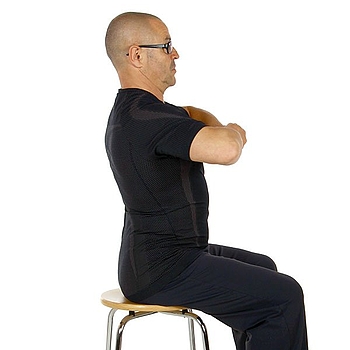 Ein Mann mit Brille trägt schwarze Kleidung. Er sitzt auf einem Hocker, seine Arme sind vor der Brust, die Hände greifen ineinander. Er zeigt eine Übung zur Mobilisation der Wirbelsäule