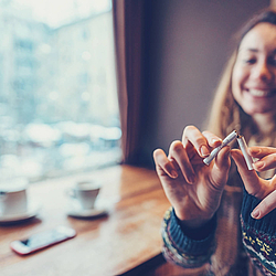 Eine Frau mit langen Haaren sitzt in einem Café. Sie zerbricht eine Zigarette und lächelt dabei.
