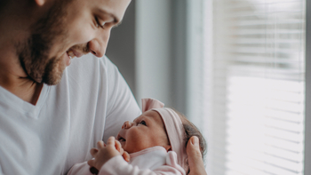 Ein junger Vater hält sein Neugeborenes im Arm. Die beiden schauen sich an und lächeln.