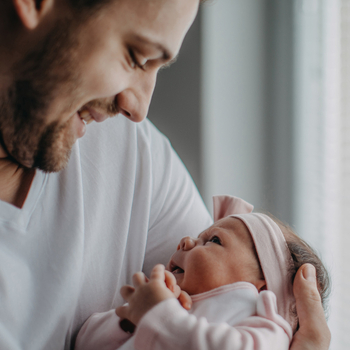 Ein junger Vater hält sein Neugeborenes im Arm. Die beiden schauen sich an und lächeln.