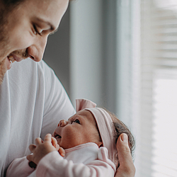 Ein junger Vater hält sein Neugeborenes im Arm. Die beiden schauen sich an und lächeln.