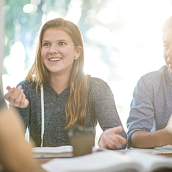 Eine junge Frau mit blonden Haaren, sitzt mit anderen jungen Leuten an einem Tisch und unterhält sich lächelnd. 