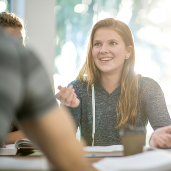 Eine junge Frau mit langen blonden Haaren, sitzt mit anderen jungen Leuten an einem Tisch und unterhält sich lächelnd. Auf dem Tisch liegen aufgeschlagene Bücher.