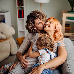 Eine junge Familie sitzt auf dem Sofa. Der Vater hat lange lockige Haare, seine Hand liegt auf dem Bauch der schwangeren Mutter. Sie hält ein kleines Mädchen im Arm.